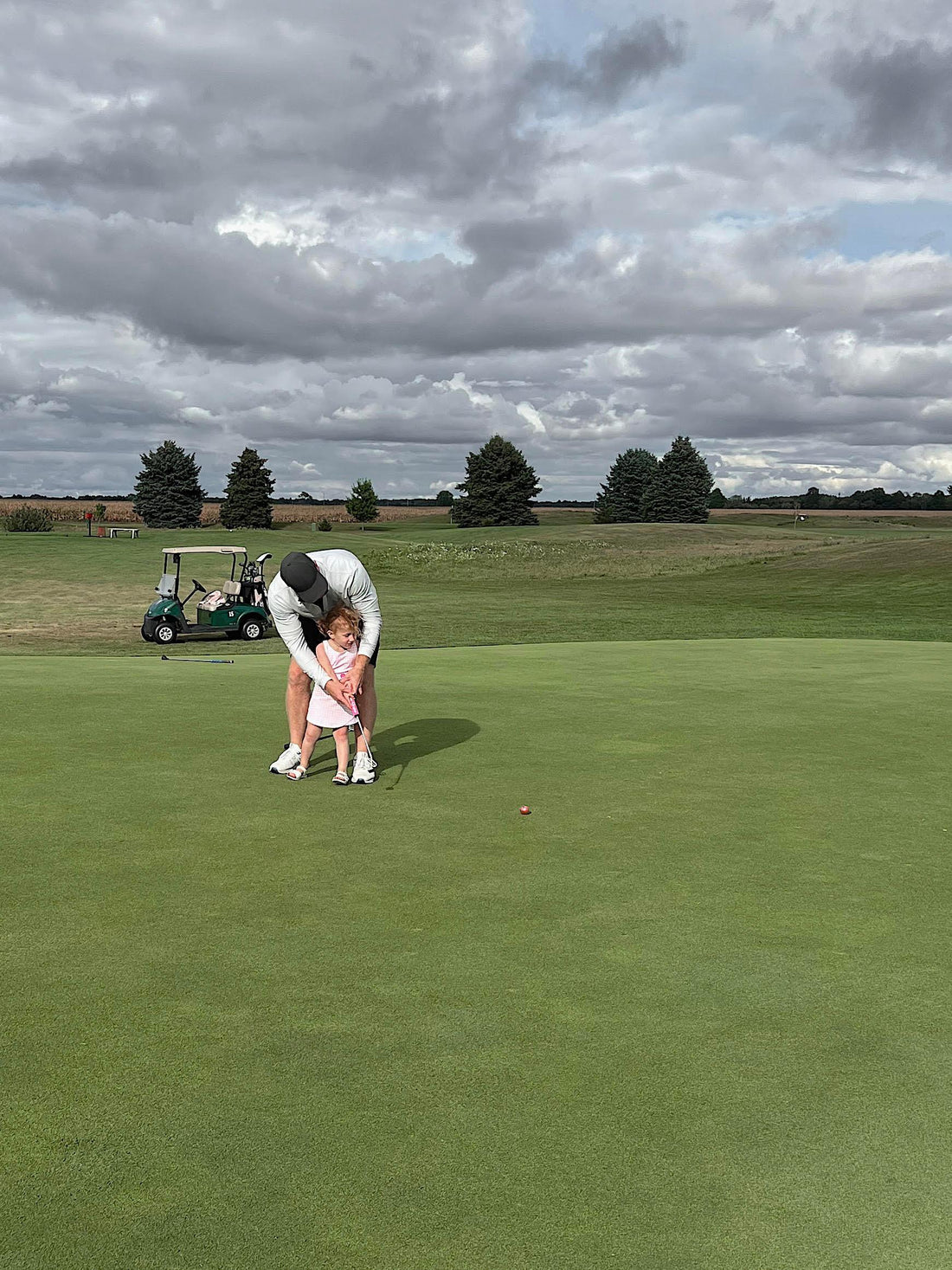 Adult teaching young girl ages 2-5 toddler golf on green, golf cart in background
