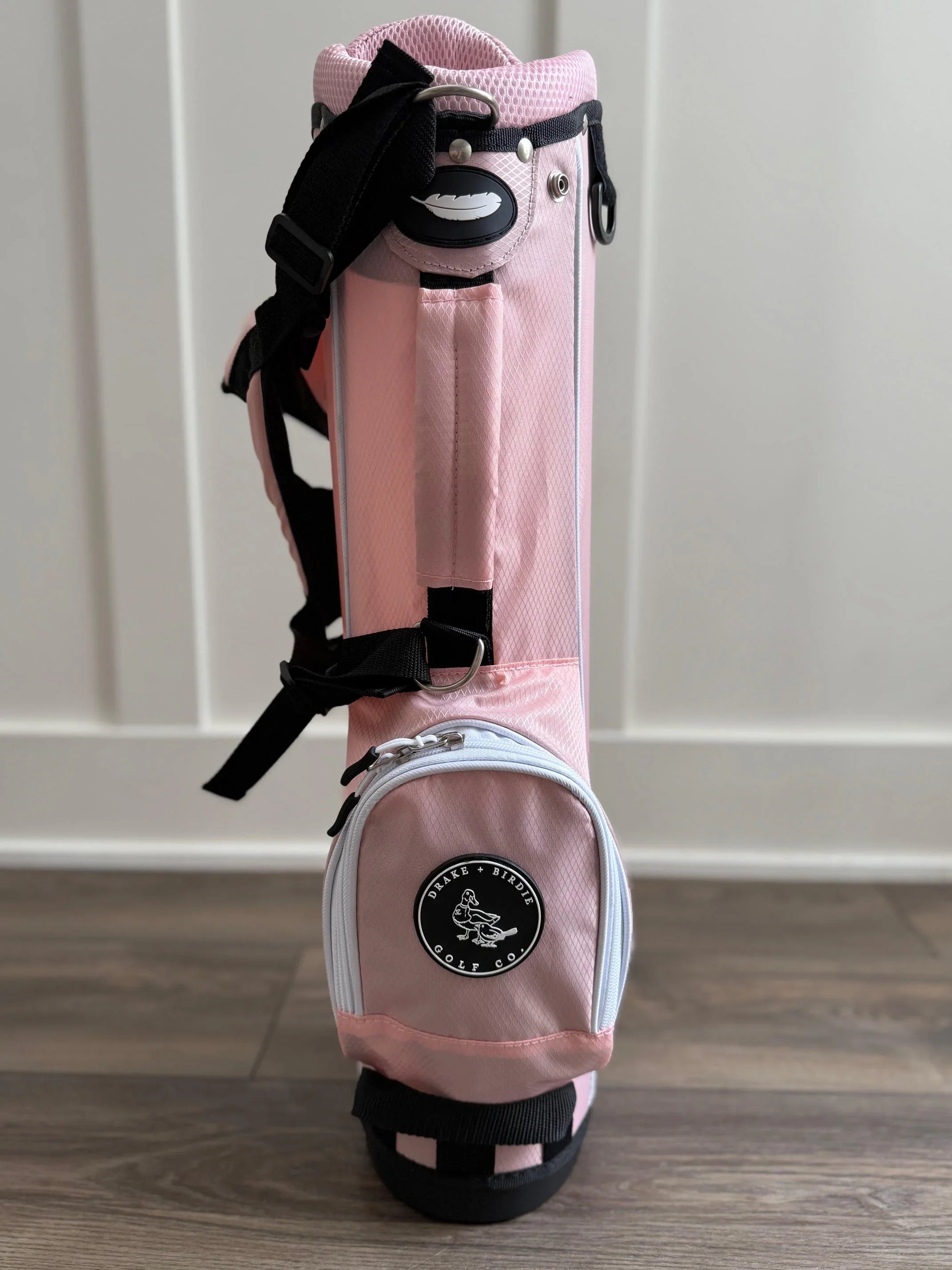 Pink toddler golf bag with black straps on a wooden floor.