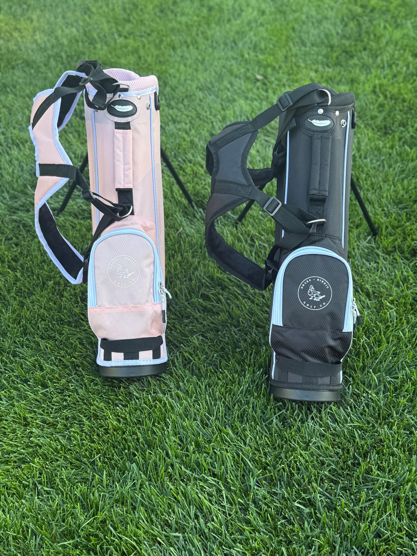 Two golf bags, one pink and one black with a logo, on grass.